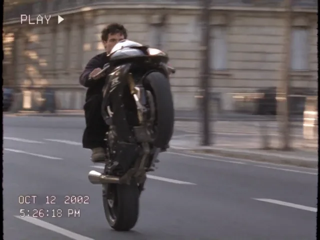 Ein Motorrad fährt auf dem Hinterrad an der Kamera vorbei, der Fahrer lehnt weit zurück, die Straße ist von Altbauten gesäumt