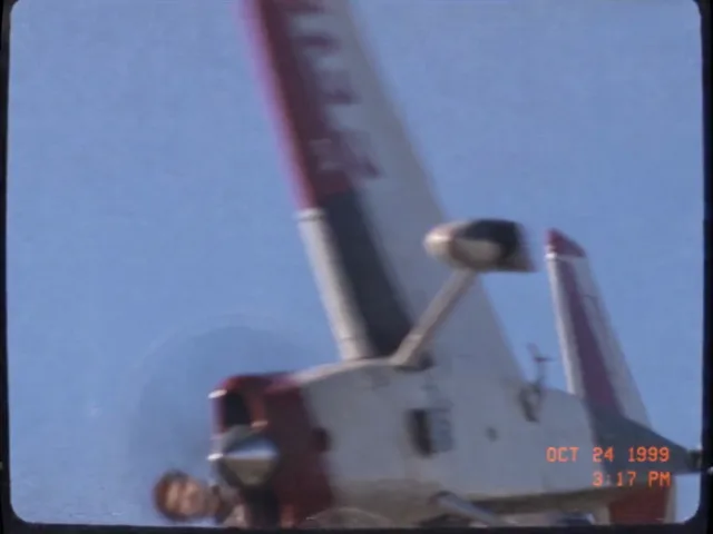 Ein rot-weißer Kunstflieger dreht sich vor blauem Himmel um die Längsachse, auf der Tragfläche steht ein Holzstuhl mit angeschnalltem Mann