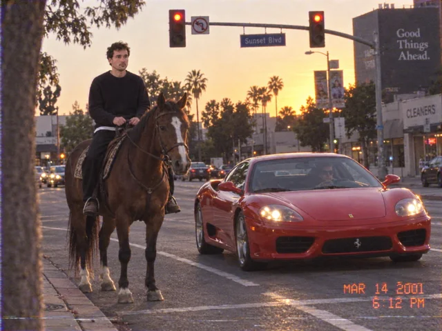 Camcorder-Standbild an einer Kreuzung im Abendlicht: links ein Reiter auf einem Pferd, rechts daneben ein roter Ferrari, beide an der roten Ampel