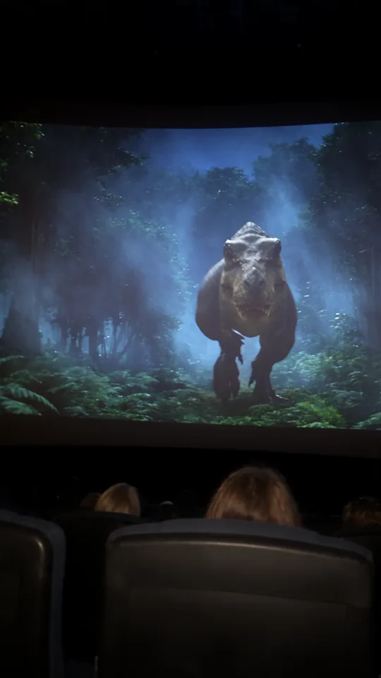 Die Kinoleinwand füllt das Bild: ein T-Rex rennt durch einen nebligen Dschungel frontal auf die Kamera zu, davor die Köpfe des Publikums