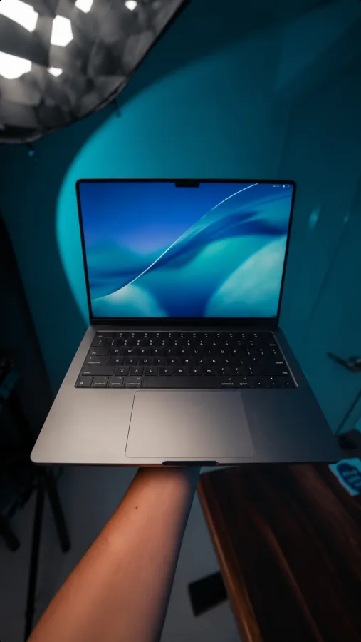 Screenshot des ersten Videoframes: eine Hand hält ein aufgeklapptes MacBook vor einer türkis beleuchteten Studiowand