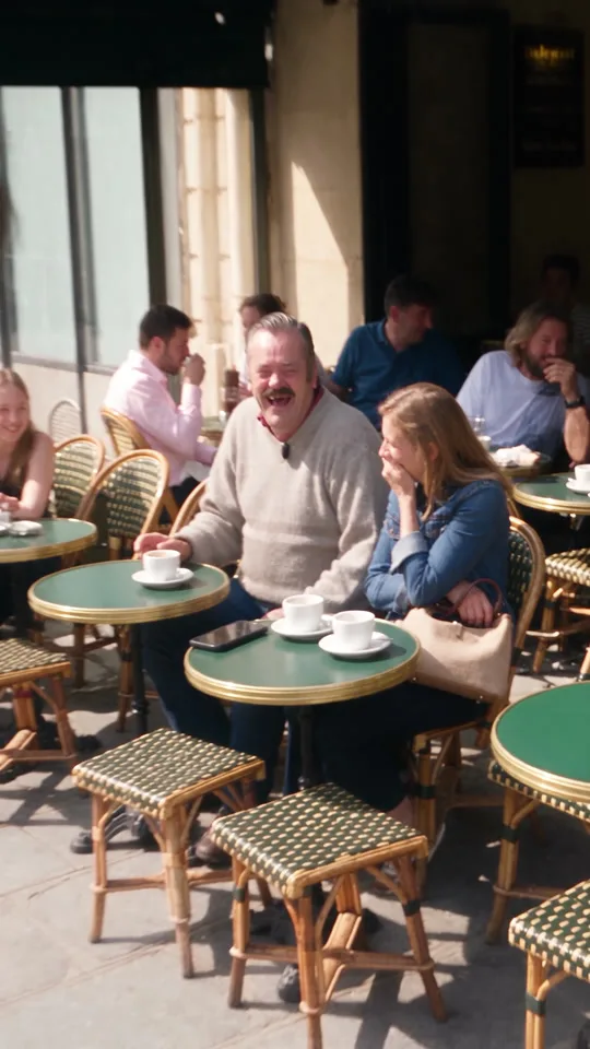 Ein Mann mit Schnurrbart lacht auf einer Pariser Caféterrasse unkontrolliert, die Gäste am Nebentisch reagieren nicht