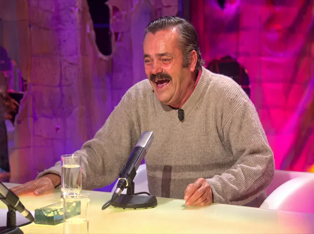 Mann mit Schnurrbart und grauem Strickpullover lacht in einem Fernsehstudio unkontrolliert